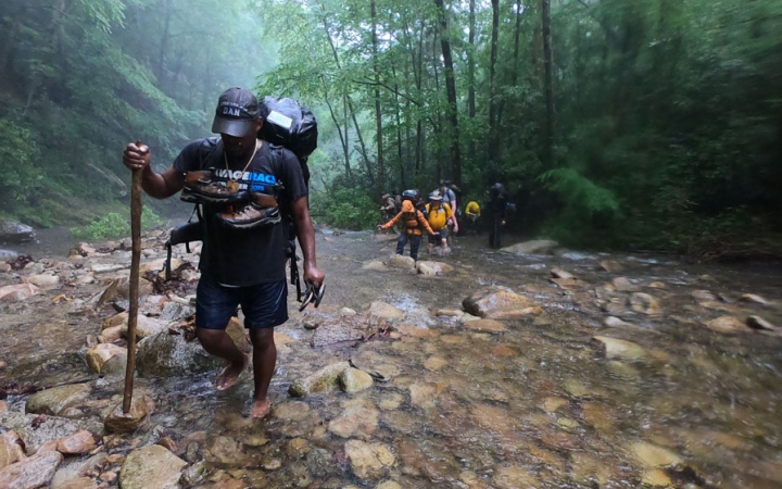 a person carrying a backpacking uses a walking stick to cross a creek. there are green trees in the background.